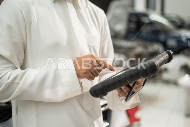 مهندس ميكانيكي محترف, استخدام التقنية والتكنولوجيا لصيانة السياره في الورشه, طلب تصليح السياره عن طريق جهاز التابلت, صورة مقربة رجل عربي سعودي خليجي يرتدي الزي السعودي التقليدي يقف في مركز صيانة السيارات