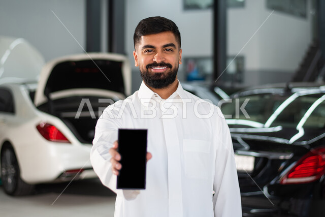 عرض شاشة سوداء فارغة عبر الهاتف المحمول ، رجل عربي خليجي سعودي يرتدي الزي السعودي التقليدي، ايماءات وجه تدل على الفرح والسرور , خدمات اصلاح و صيانة السيارات ، مهندس ميكانيكي ، ورشة صيانة