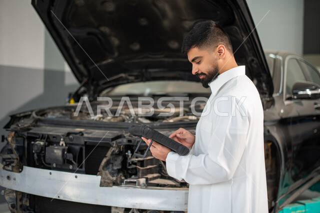طلب تصليح السياره عن طريق جهاز التابلت, استخدام التقنيه والتكنولوجيا لصيانة السياره في الورشه, رجل عربي سعودي خليجي يرتدي الزي السعودي التقليدي يقف في مركز صيانة السيارات, مهندس ميكانيكي محترف