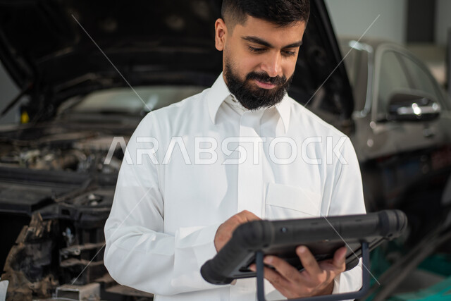 استخدام التكنولوجيا والتقنية لصيانة السياره في الورشه, طلب تصليح السياره عن طريق جهاز التابلت, رجل عربي سعودي خليجي يرتدي الزي السعودي التقليدي يقف في مركز صيانة السيارات, مهندس ميكانيكي محترف