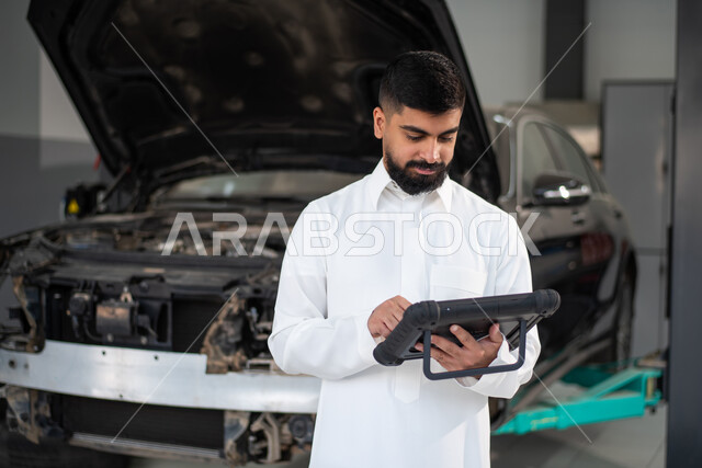 استخدام التقنيه والتكنولوجيا لصيانة السياره في الورشه, طلب تصليح السياره عن طريق جهاز التابلت, رجل عربي سعودي خليجي يرتدي الزي السعودي التقليدي يقف في مركز صيانة السيارات, مهندس ميكانيكي محترف