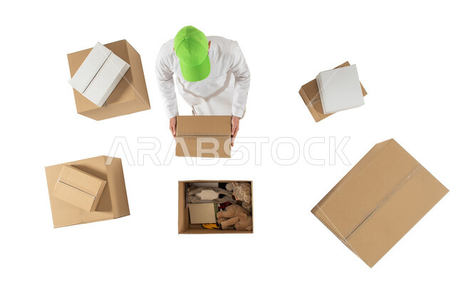 امين مستودع يرتدي قبعة خضراء ويحمل صندوق كرتوني، تعبئة وتغليف صناديق الطلبات، صورة علوية عامودية لرجل خليجي سعودي، مندوب توصيل الطلبات والبضائع للزبائن، التشيك على الحجوزات، خدمات الشحن والتوصيل، خلفية بيضاء