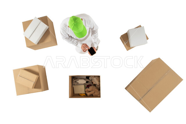 مندوب توصيل الطلبات والبضائع للزبائن يستخدم الهاتف المحمول، امين مستودع يرتدي قبعة خضراء، صورة علوية عامودية لرجل سعودي، التسويق الالكتروني الذكي، تغلفة صناديق الطلبات، التشيك على الحجوزات، خدمات الشحن والتوصيل، خلفية بيضاء