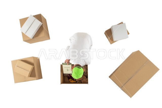 امين مستودع يقوم بتعبئة وتغليف صناديق الطلبات، صورة علوية عامودية لرجل خليجي سعودي، مندوب توصيل الطلبات والبضائع للزبائن يرتدي قبعة خضراء، التشيك على الحجوزات، خدمات الشحن والتوصيل، خلفية بيضاء