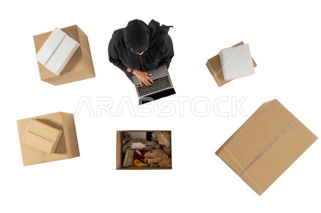 امينة مستودع تستخدم الحاسوب المحمول، صورة علوية عامودية لامرأة سعودية، مندوبة توصيل الطلبات والبضائع للزبائن، التسويق الالكتروني الذكي، تغليف صناديق الطلبات، التشيك على الحجوزات، خدمات الشحن والتوصيل، خلفية بيضاء