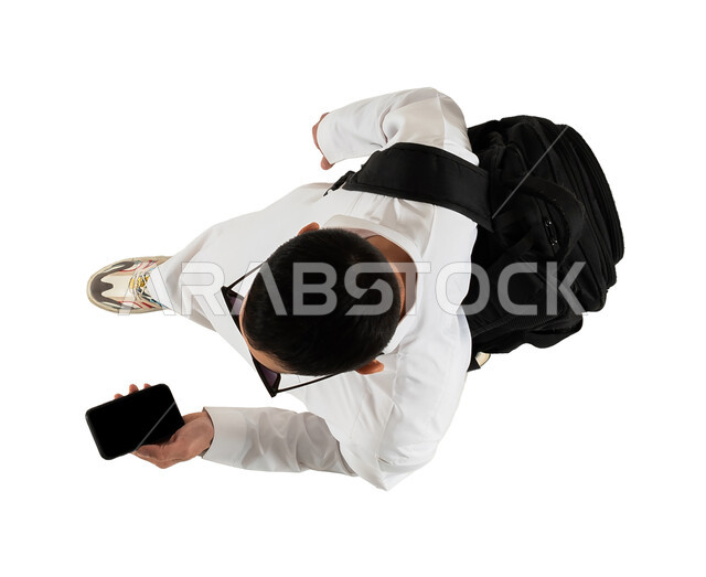 شاب يرتدي النظارات الشمسية ويستخدم الهاتف المحمول، صورة علوية عامودية لرجل عربي خليجي سعودي يحمل حقيبة الظهر الشخصية ويستعد لقضاء الاجازة الصيفية، تصفح مواقع وتطبيقات السياحة والسفر، الذهاب في رحلة سياحية ممتعة، خلفية بيضاء