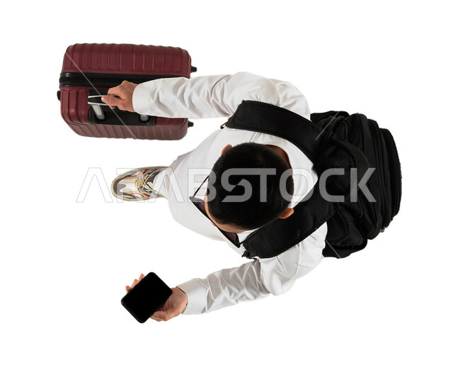 شاب يسمك بيده حقيبة السفر، الاستعداد للسفر والسياحة، اجازة صيفية سياحية، صورة علوية عامودية لرجل عربي خليجي سعودي يرتدي النظارات الشمسية وحقيبة الظهر الشخصية، تصفح مواقع وتطبيقات السياحة والسفر من خلال الهاتف المحمول، خلفية بيضاء