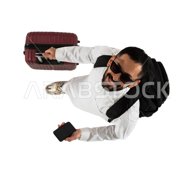 الاستعداد للسفر والسياحة، اجازة صيفية سياحية، صورة علوية عامودية لرجل عربي خليجي سعودي يرتدي النظارات الشمسية وحقيبة الظهر الشخصية، شاب ينظر الى الاعلى ويسمك بيده حقيبة السفر، تصفح مواقع وتطبيقات السياحة والسفر من خلال الهاتف المحمول، خلفية بيضاء