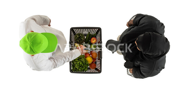 مندوب توصيل طلبات يرتدي قبعة خضراء، تسليم واستلام الطلبات، سلة تسوق مليئة بالخضروات والفواكه، صورة علوية عامودية لامرأة ورجل عربيان خليجيان سعوديان، توصيل طلبات الديلفري، خدمات التوصيل السريع للطلبات المنزلية، خلفية بيضاء