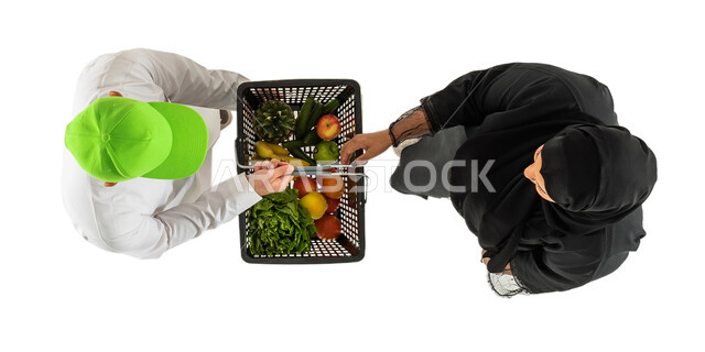 مندوب توصيل طلبات يرتدي قبعة خضراء ويحمل سلة تسوق مليئة بالخضروات والفواكه، صورة علوية عامودية لرجل وامرأة سعوديان عربيان خليجيان، توصيل طلبات الديلفري، تسليم واستلام الطلبات، خدمات التوصيل السريع للطلبات المنزلية، خلفية بيضاء