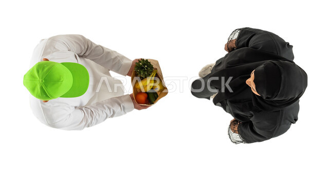 تسليم واستلام الطلبات، مندوب توصيل طلبات يرتدي قبعة خضراء ويحمل كيس الفواكه والخضروات، صورة علوية عامودية لامرأة ورجل عربيان خليجيان سعوديان، توصيل طلبات الديلفري، خدمات التوصيل السريع للطلبات المنزلية، خلفية بيضاء