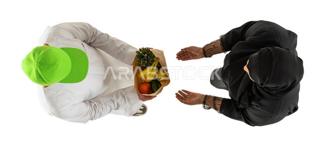 مندوب توصيل طلبات يرتدي قبعة خضراء ويحمل كيس الفواكه والخضروات، صورة علوية عامودية لامرأة ورجل عربيان خليجيان سعوديان، توصيل طلبات الديلفري، تسليم واستلام الطلبات، خدمات التوصيل السريع للطلبات المنزلية، خلفية بيضاء