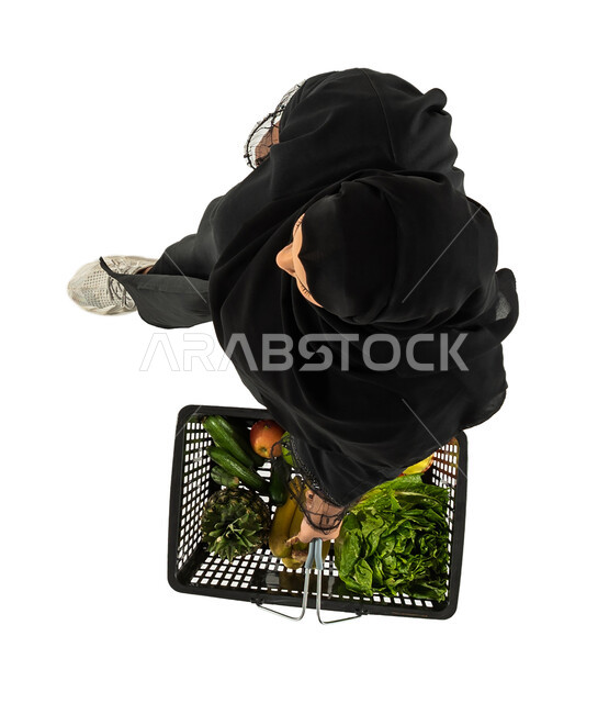 شراء الاحتياجات المنزلية، صورة علوية عامودية لامرأة عربية خليجية سعودية تحمل سلة التسوق، السلع والمواد الغذائية المختلفة، مستلزمات المستهلك، خلفية بيضاء