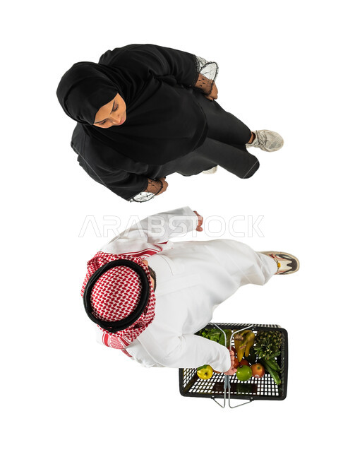 رجل يرتدي الزي السعودي التقليدي ويحمل سلة التسوق، صورة علوية عامودية لزوجان عربيان خليجيان سعوديان يقومان بشراء الاحتياجات المنزلية، السلع والمواد الغذائية المختلفة، مستلزمات المستهلك، خلفية بيضاء