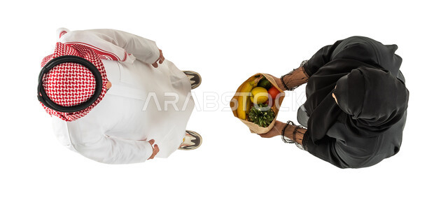 امرأة خليجية سعودية تحمل بيدها كيس من الفواكه والخضروات، صورة علوية عامودية لامرأة ورجل عربيان خليجيان سعوديان، توصيل طلبات الديلفري، تسليم واستلام الطلبات، خدمات التوصيل السريع للطلبات المنزلية، خلفية بيضاء