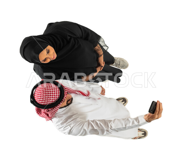 التقاط صور تذكارية من خلال الجوال، صورة علوية عامودية لزوجان عربيان سعوديان خليجيان يستخدمان الهاتف المحمول، اجراء مكالمة فيديو، بث مباشر، خلفية بيضاء
