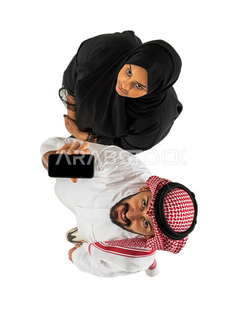 رجل يمسك هاتف محمول بشاشة سوداء فارغة، صورة علوية عامودية لزوجان عربيان خليجيان سعوديان، عرض خدمات وتطبيقات الهاتف المحمول، التسوق الإلكتروني، ايماءات وجه سعيدة، خلفية بيضاء
