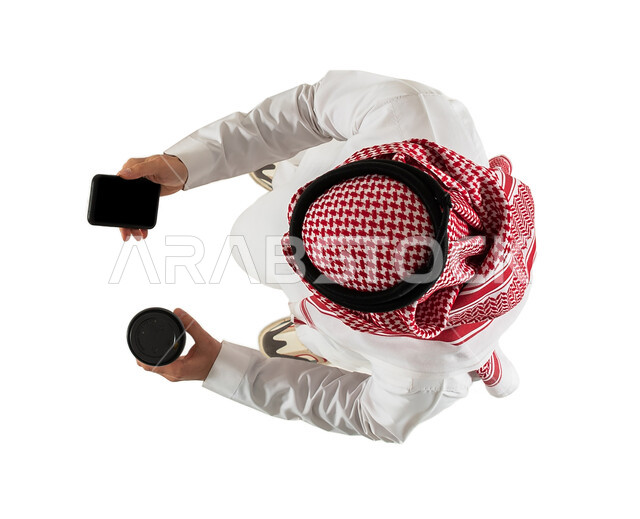 الاستمتاع بالمشروبات ساخنة، صورة علوية عامودية لرجل عربي خليجي سعودي يستخدم الهاتف المحمول وبيده كوب، التسوق الالكتروني وطلبات اونلاين، عملية شراء الكترونية، خدمات التوصيل السريع للمشروبات الساخنة، تصفح المواقع الالكترونية، خلفية بيضاء