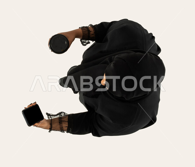 التسوق الالكتروني وطلبات اونلاين، الاستمتاع بالمشروبات ساخنة، صورة علوية عامودية لامرأة عربية خليجية سعودية تستخدم الهاتف المحمول وبيدها كوب قهوة، عملية شراء الكترونية، خدمات التوصيل السريع للمشروبات الساخنة، تصفح المواقع الالكترونية، خلفية بيضاء