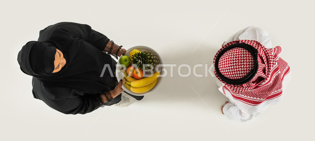 اكرام الضيف وتقديم طبق الفواكه الطازجة، حسن الضيافة والاستقبال، صورة علوية عامودية لرجل وامرأة عربيان خليجيان سعوديان، ارتداء الزي السعودي التقليدي، خلفية بيضاء