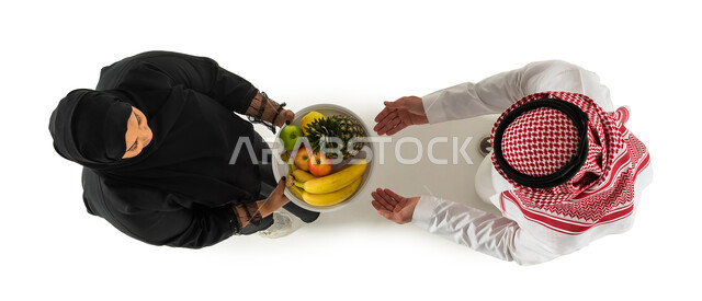 اكرام الضيف وتقديم طبق الفواكه الطازجة، صورة علوية عامودية لرجل وامرأة عربيان خليجيان سعوديان، ارتداء الزي السعودي التقليدي، حسن الضيافة والاستقبال، خلفية بيضاء