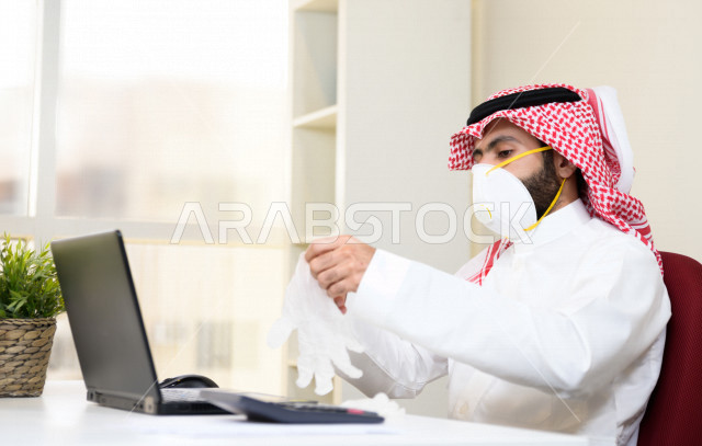 رجل اعمال عربي سعودي خليجي يرتدي الكمامة والقفازات لضبط انتشار العدوى ، يجلس امام الحاسوب المحمول لانجاز العمل، ايماءات يدين مختلفة ، اجتماعات اونلاين ، العمل عن بعد، التسويق الالكتروني