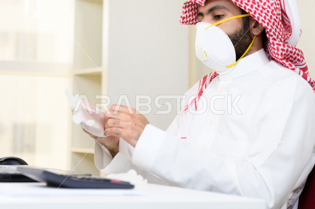 رجل اعمال عربي سعودي خليجي يرتدي الكمامة والقفازات لضبط انتشار العدوى ، يجلس امام الحاسوب المحمول لانجاز العمل، ايماءات يدين مختلفة ، اجتماعات اونلاين ، العمل عن بعد، التسويق الالكتروني