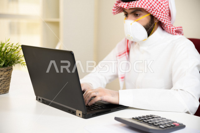 رجل اعمال عربي سعودي خليجي يرتدي الكمامة والقفازات لضبط انتشار العدوى ، استخدام الحاسوب المحمول لانجاز العمل ، القيام بالعمليات المحاسبية ، العمل عن بعد ، اجتماعات اونلاين ، التسويق الالكتروني