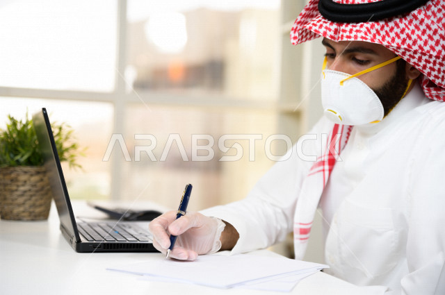 رجل اعمال عربي سعودي خليجي يرتدي الكمامة والقفازات لضبط انتشار العدوى  ، يقوم بتدوين الملاحظات على الورق  ، استخدام الحاسوب المحمول لانجاز العمل، العمل عن بعد، اجتماعات اونلاين ، التسويق الالكتروني