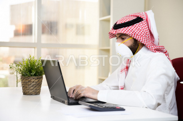 رجل اعمال عربي سعودي خليجي يرتدي الكمامة والقفازات لضبط انتشار العدوى ، استخدام الحاسوب المحمول لانجاز العمل ، القيام بالعمليات المحاسبية ، العمل عن بعد ، اجتماعات اونلاين ، التسويق الالكتروني