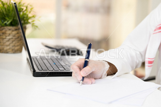 رجل اعمال عربي سعودي خليجي يرتدي الكمامة والقفازات لضبط انتشار العدوى ، يقوم بتدوين الملاحظات على الورق ، استخدام الحاسوب المحمول لانجاز العمل، العمل عن بعد، اجتماعات اونلاين ، التسويق الالكتروني