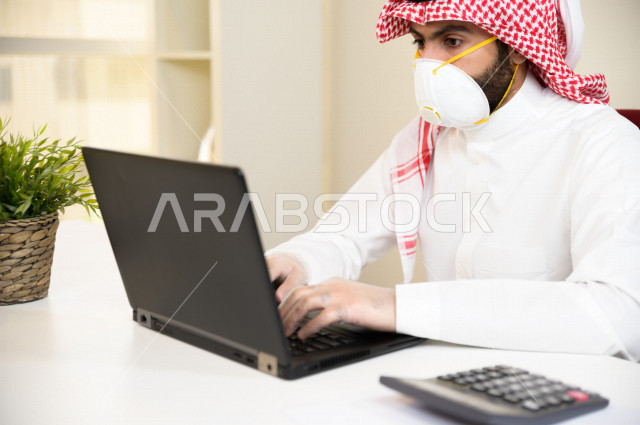 رجل اعمال عربي سعودي خليجي يرتدي الكمامة والقفازات لضبط انتشار العدوى ، استخدام الحاسوب المحمول لانجاز العمل ، القيام بالعمليات المحاسبية ، العمل عن بعد ، اجتماعات اونلاين ، التسويق الالكتروني