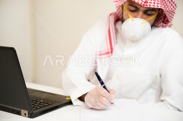 رجل اعمال عربي سعودي خليجي يرتدي الكمامة والقفازات لضبط انتشار العدوى  ، يقوم بتدوين الملاحظات على الورق  ، استخدام الحاسوب المحمول لانجاز العمل، العمل عن بعد، اجتماعات اونلاين ، التسويق الالكتروني