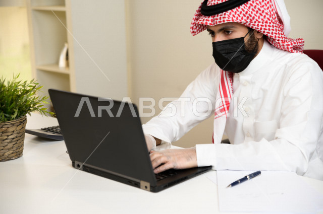 رجل اعمال عربي سعودي خليجي يرتدي الكمامة والقفازات لضبط انتشار العدوى ، استخدام الحاسوب المحمول لانجاز العمل ، القيام بالعمليات المحاسبية ، العمل عن بعد ، اجتماعات اونلاين ، التسويق الالكتروني