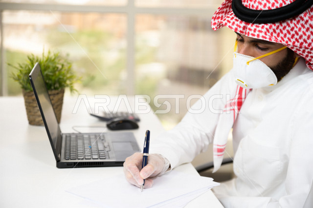 رجل اعمال عربي سعودي خليجي يرتدي الكمامة والقفازات لضبط انتشار العدوى  ، يقوم بتدوين الملاحظات على الورق  ، استخدام الحاسوب المحمول لانجاز العمل، العمل عن بعد، اجتماعات اونلاين ، التسويق الالكتروني