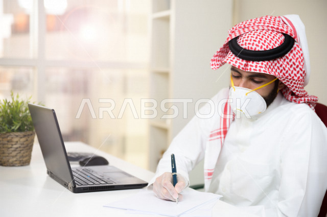 رجل اعمال عربي سعودي خليجي يرتدي الكمامة والقفازات لضبط انتشار العدوى  ، يقوم بتدوين الملاحظات على الورق  ، استخدام الحاسوب المحمول لانجاز العمل، العمل عن بعد، اجتماعات اونلاين ، التسويق الالكتروني