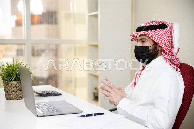 رجل اعمال عربي سعودي خليجي يرتدي الكمامة والقفازات لضبط انتشار العدوى ، يجلس امام الحاسوب المحمول لانجاز العمل، ايماءات يدين مختلفة ، اجتماعات اونلاين ، العمل عن بعد، التسويق الالكتروني