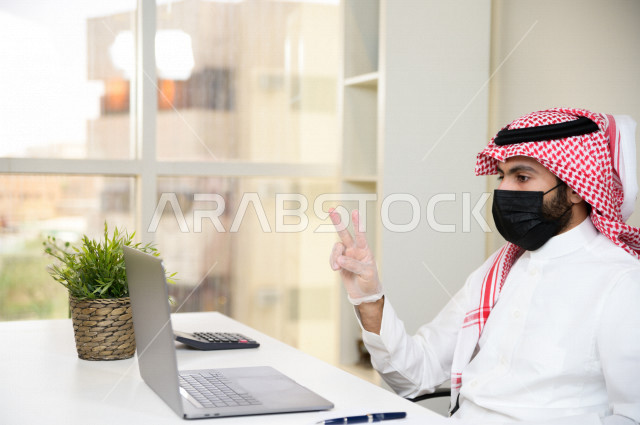 رجل اعمال عربي سعودي خليجي يرتدي الكمامة والقفازات لضبط انتشار العدوى ، يجلس امام الحاسوب المحمول لانجاز العمل، ايماءات يدين مختلفة ، اجتماعات اونلاين ، العمل عن بعد، التسويق الالكتروني