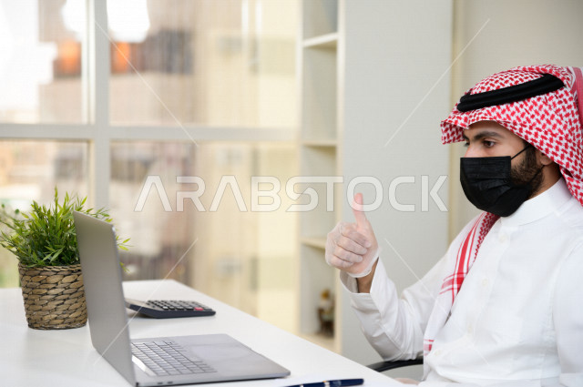 رجل اعمال عربي سعودي خليجي يرتدي الكمامة والقفازات لضبط انتشار العدوى ، يجلس امام الحاسوب المحمول لانجاز العمل، ايماءات يدين مختلفة ، اجتماعات اونلاين ، العمل عن بعد، التسويق الالكتروني