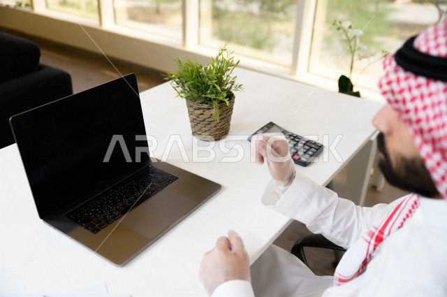 رجل اعمال عربي سعودي خليجي يرتدي القفازات لضبط انتشار العدوى ، استخدام الحاسوب المحمول لانجاز العمل ، العمل عن بعد من المنزل ، اجتماعات اونلاين ، التسويق الالكتروني