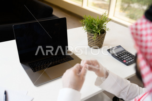 رجل اعمال عربي سعودي خليجي يرتدي القفازات لضبط انتشار العدوى ، استخدام الحاسوب المحمول لانجاز العمل ، العمل عن بعد من المنزل ، اجتماعات اونلاين ، التسويق الالكتروني