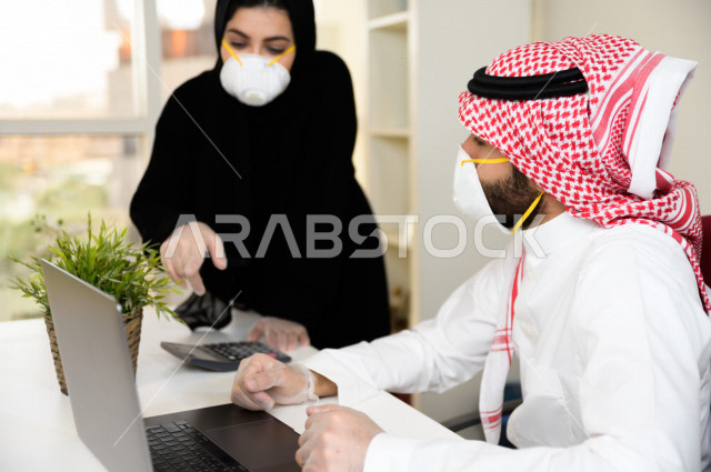 رجل اعمال عربي سعودي خليجي يرتدي الكمامة والقفازات لضبط انتشار العدوى ،بجانبه سكرتيرة خليجية سعودية ترتدي الكمامة والقفازات، استخدام الحاسوب المحمول لانجاز العمل ، متابعة الاعمال الادارية ، اتباع اجراءات الوقاية والسلامة من فايروس كورونا