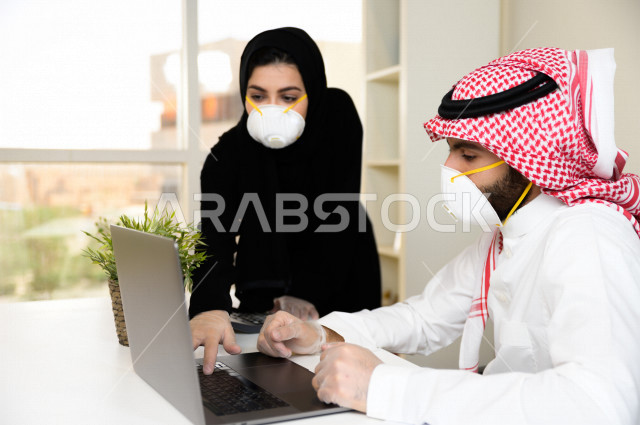 رجل اعمال عربي سعودي خليجي يرتدي الكمامة والقفازات لضبط انتشار العدوى ،بجانبه سكرتيرة خليجية سعودية ترتدي الكمامة، استخدام الحاسوب المحمول لانجاز العمل ، متابعة الاعمال الادارية ، اتباع اجراءات الوقاية والسلامة من فايروس كورونا