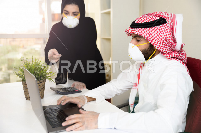 رجل اعمال عربي سعودي خليجي يرتدي الكمامة والقفازات لضبط انتشار العدوى ،بجانبه سكرتيرة خليجية سعودية ترتدي الكمامة، استخدام الحاسوب المحمول لانجاز العمل ، متابعة الاعمال الادارية ، اتباع اجراءات الوقاية والسلامة من فايروس كورونا