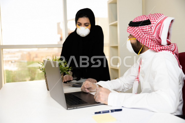 رجل اعمال عربي سعودي خليجي يرتدي الكمامة والقفازات لضبط انتشار العدوى ،بجانبه سكرتيرة خليجية سعودية ترتدي الكمامة، استخدام الحاسوب المحمول لانجاز العمل ، متابعة الاعمال الادارية ، اتباع اجراءات الوقاية والسلامة من فايروس كورونا