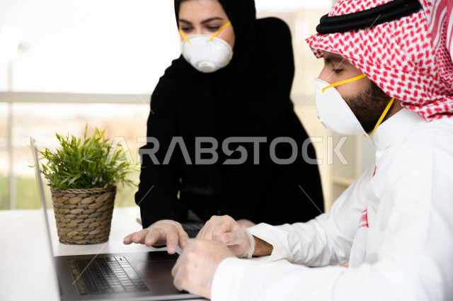 رجل اعمال عربي سعودي خليجي يرتدي الكمامة والقفازات لضبط انتشار العدوى ،بجانبه سكرتيرة خليجية سعودية ترتدي الكمامة، استخدام الحاسوب المحمول لانجاز العمل ، متابعة الاعمال الادارية ، اتباع اجراءات الوقاية والسلامة من فايروس كورونا