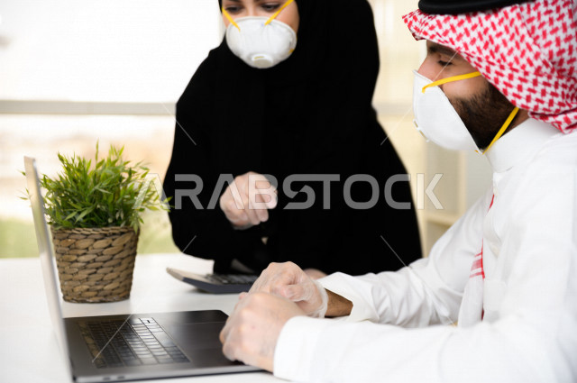 رجل اعمال عربي سعودي خليجي يرتدي الكمامة والقفازات لضبط انتشار العدوى ،بجانبه سكرتيرة خليجية سعودية ترتدي الكمامة والقفازات، استخدام الحاسوب المحمول لانجاز العمل ، متابعة الاعمال الادارية ، اتباع اجراءات الوقاية والسلامة من فايروس كورونا