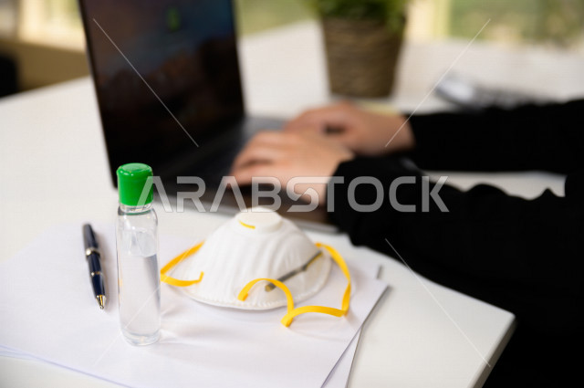 A Saudi Arabian Gulf businesswoman sitting in front of a laptop to complete tasks, with hand sanitizer and a mask, following safety and prevention measures, doing administrative work remotely to avoid the spread of infection