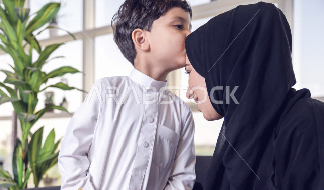 A Saudi Arabian Gulf child kissing his mother's head on the morning of Eid with gestures indicating happiness and joy, wearing traditional Saudi dress, Eidiya, Gergean, celebrations and manifestations of Eid, spending happy family times on Eid