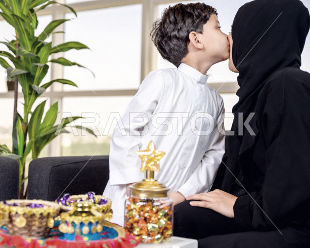 طفل عربي خليجي سعودي يقوم بتقبيل رأس والدته صباح يوم العيد بإيماءات تدل على السعادة و الفرح ، ارتداء اللباس السعودي التقليدي ، العيدية ، القرقيعان ، احتفالات و مظاهر العيد ، قضاء أوقات عائلية سعيدة في العيد